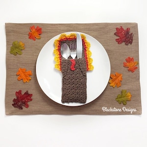 May include: A crocheted turkey place card holder with a brown body, red head, yellow and orange feathers, and a white plate with a fork and spoon inside. The place card holder is on a brown placemat with fall leaves.