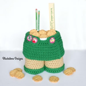 May include: A green crocheted Girl Scout uniform shaped like a container filled with Girl Scout cookies. The uniform has a tan sash and two badges. A ruler with the text "G.I.R.L.S RULE! Go-Getters" is standing upright in the container. Two green pencils are also in the container. There are several Girl Scout cookies scattered on a white surface.