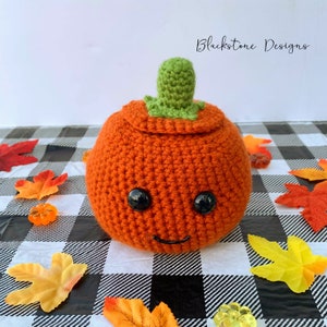 May include: A crocheted orange pumpkin with a green stem and black eyes. The pumpkin has a smiling mouth and is sitting on a black and white checked surface.