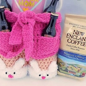 May include: A pink crocheted gift basket with bunny slippers, holding miniature bottles of Bailey's Irish Cream. A bag of New England Coffee, Blueberry Cobbler flavor, is next to the basket. The text "MOM" is visible.