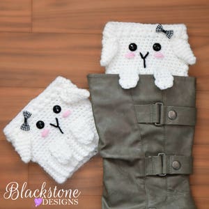 Pode incluir: Duas punhos de bota de cordeiro de crochê branco com olhos de botão preto, bochechas rosa e um laço preto. Um punho está dentro de uma bota cinza, e o outro está ao lado. A imagem inclui o texto "Blackstone Designs".