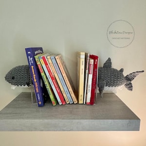 Könnte beinhalten: Zwei graue, gehäkelte Hai-Buchstützen halten eine Reihe bunter Kinderbücher auf einem Holzregal. Die Buchstützen stehen sich gegenüber und haben weiße Bäuche. Die Bücher sind hauptsächlich rot und haben Titel wie "Runaway Ralph" und "Beezus and Ramona".