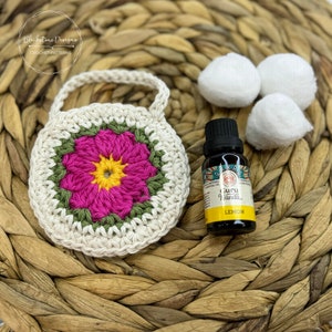 May include: A crocheted flower-shaped pouch with a handle, made in white, green, pink, and yellow yarn. The pouch is sitting on a woven wicker surface with a small bottle of lemon essential oil and cotton balls.