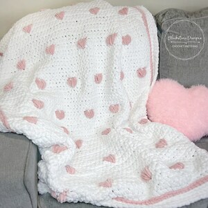 May include: White crochet blanket with pink heart-shaped embellishments. The blanket is folded and draped over a gray couch with a pink heart-shaped pillow.