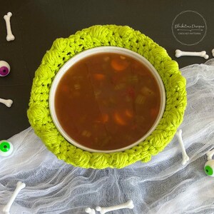 以下が含まれることがあります： 明るい緑色の編み物で囲まれた、スープが入った白いボウル。スープには人参とセロリが見えます。シーンは白い骨の形をした装飾と白いガーゼで飾られています。