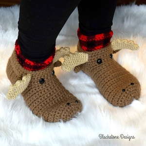 Puede incluir: Un par de pantuflas de reno de color marrón hechas a crochet con ojos negros y puños de cuadros rojos y negros. Las pantuflas se llevan sobre una superficie blanca y esponjosa.