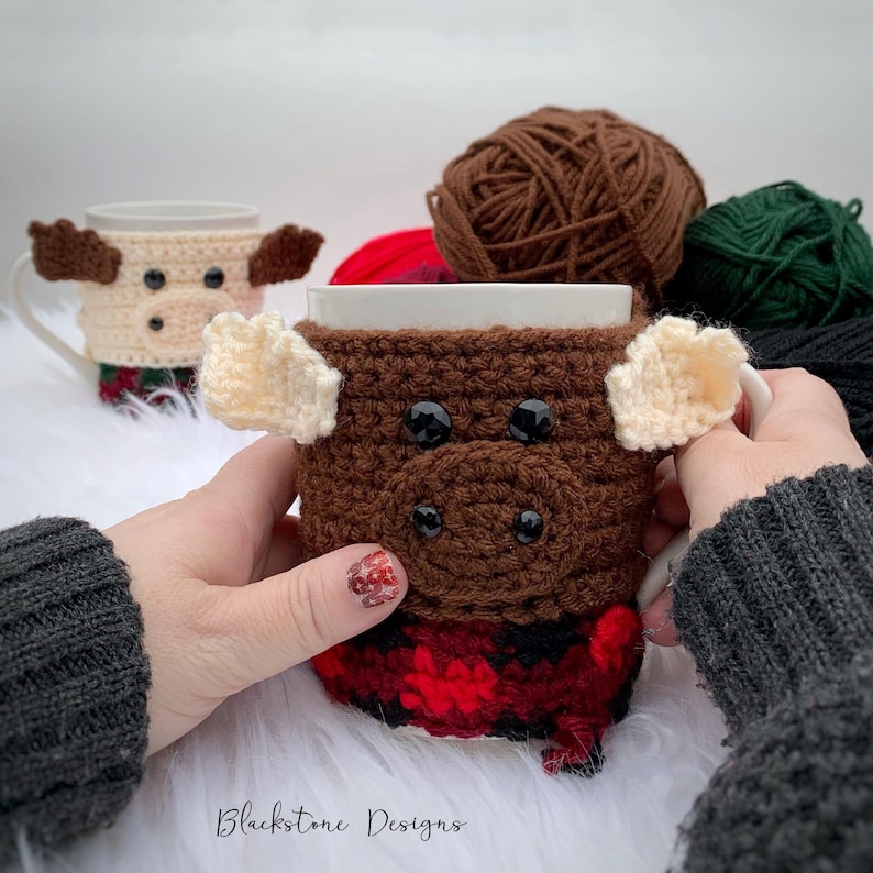 May include: A brown crocheted mug cozy shaped like a Highland cow, with cream-colored horns and black button eyes. The bottom features a red and black plaid pattern. A second mug with a similar design is in the background. The text "Blackstone Designs" is visible.