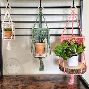 May include: Three macrame plant hangers with wooden shelves, one in white, one in green, and one in pink. Each hanger has a small potted plant on the shelf.