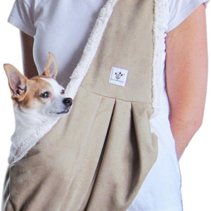 May include: A tan-colored dog carrier with a white sherpa lining. The carrier is worn by a person and has a small dog inside. The carrier has a label that says "Paw & Peace".