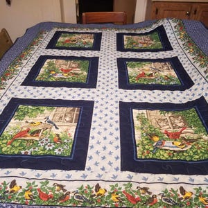 May include: A large, quilted blanket with a bird and floral design. The quilt features a central cross pattern with square panels depicting birds and cats. The border has a repeating pattern of birds and flowers, with a blue and white background.