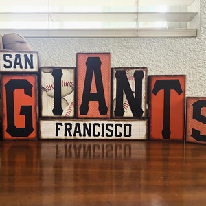 May include: Wooden block sign spelling out "SAN FRANCISCO GIANTS" in orange and black lettering. The "I" in "GIANTS" features a baseball design. A baseball and glove are visible in the background.