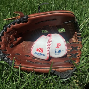 May include: A white baseball with red stitching and the text "#28 Rawlings" and "JP" in a brown leather baseball glove. The glove and ball are on a bed of green grass.