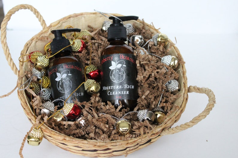 K&ouml;nnte beinhalten: Zwei braune Flaschen Handseife mit schwarzen Etiketten in einem Weidenkorb gef&uuml;llt mit braunen Papierschnitzeln und goldenen und silbernen Gl&ouml;ckchen. Die Etiketten lauten "Blueberry Botanics" und "Moisture-Rich Cleanser".