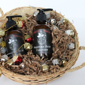 K&ouml;nnte beinhalten: Zwei braune Flaschen Handseife mit schwarzen Etiketten in einem Weidenkorb gef&uuml;llt mit braunen Papierschnitzeln und goldenen und silbernen Gl&ouml;ckchen. Die Etiketten lauten "Blueberry Botanics" und "Moisture-Rich Cleanser".