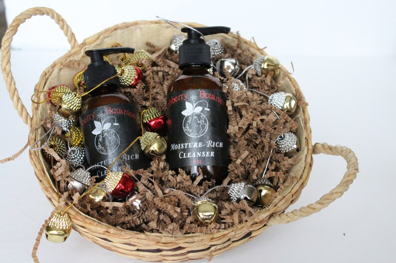 K&ouml;nnte beinhalten: Zwei braune Flaschen Handseife mit schwarzen Etiketten in einem Weidenkorb gef&uuml;llt mit braunen Papierschnitzeln und goldenen und silbernen Gl&ouml;ckchen. Die Etiketten lauten "Blueberry Botanics" und "Moisture-Rich Cleanser".