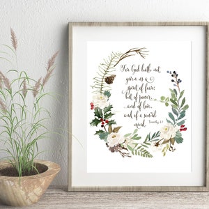 May include: A framed art print with a floral wreath design, including white flowers, green leaves, and red berries. The print displays the text "For God hath not given us a spirit of fear..." The frame is a light brown colour.