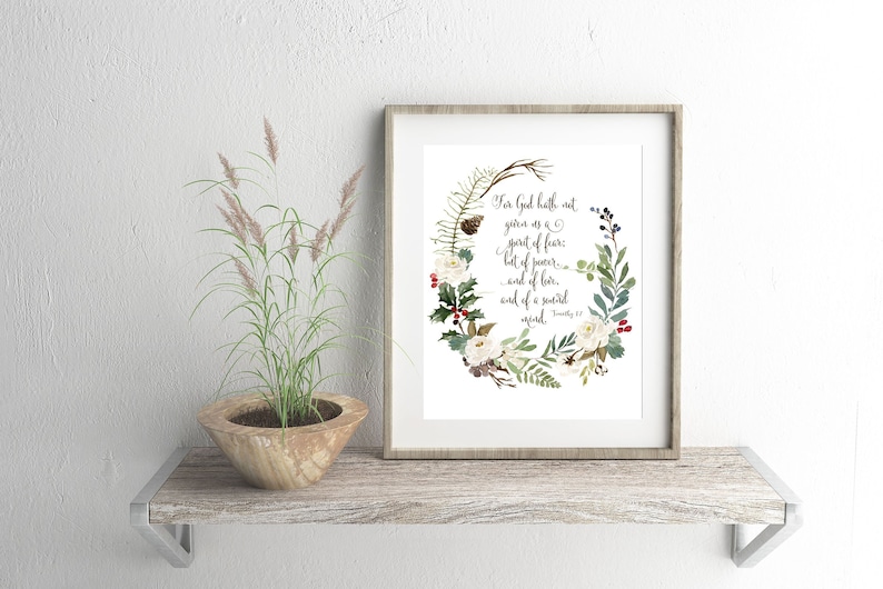 May include: A framed art print with a floral wreath design, including white flowers, green leaves, and red berries. The print displays the text "For God hath not given us a spirit of fear..." The frame is a light brown colour.
