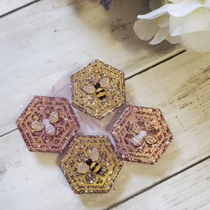 May include: Four hexagonal resin ornaments with a glitter finish. Each ornament features a bee with black and yellow stripes and a pink honeycomb design.