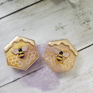May include: Two gold hexagonal resin earrings with a bee and honeycomb design. The bee is black and yellow with a white rhinestone body.