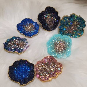 May include: Six different resin coasters with a geode design. The coasters are in various colors, including blue, pink, and teal, and have a glittery finish.