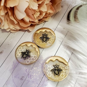 May include: Three round, gold-toned resin coasters with a white honeycomb design and a gold bee charm. The coasters are on a white surface.