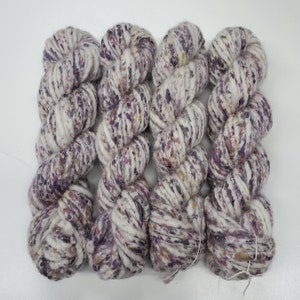 May include: Four skeins of chunky yarn in a square arrangement. The yarn is a mix of white, purple, and flecks of yellow and pink. The yarn is thick and twisted, suggesting it is for knitting or crocheting projects.