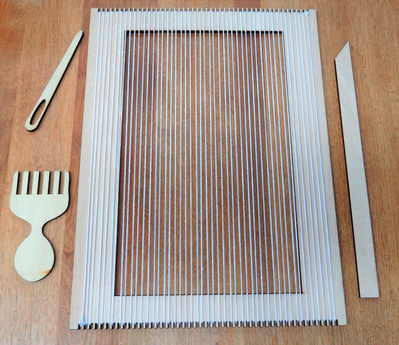 May include: A wooden loom with white string warp threads, a wooden comb, a wooden needle, and a wooden dowel. The loom is on a wooden surface.