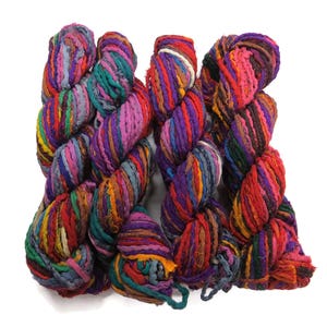 Pode incluir: Um novelo de fio colorido, fiado à mão a partir de sobras de tecido recicladas. O fio é uma mistura de roxo, rosa, vermelho, laranja, amarelo, verde, azul e castanho.