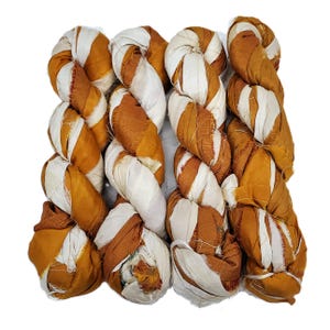 May include: Four skeins of hand-dyed silk ribbon yarn arranged side by side. Each skein features a twisted, braided texture with alternating strips of solid mustard yellow and off-white fabric, showcasing frayed edges and a soft, matte finish.