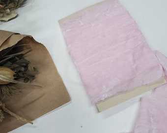 May include: A close-up of a pale pink velvet ribbon. The ribbon is soft and has a subtle sheen. It is displayed on a white surface.