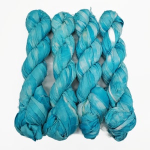 Puede incluir: Cuatro ovillos de hilo de cinta de seda azul turquesa. El hilo está retorcido y tiene un patrón de mármol blanco y turquesa.