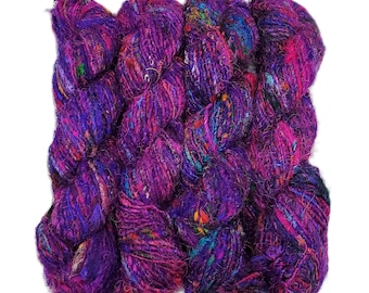 Könnte beinhalten: Ein Garnstrang mit strukturierter Oberfläche in einer lebendigen Palette aus tiefem Violett, Magenta und Purpur, durchsetzt mit bunten Sprenkeln in Blau, Orange und Grün. Das Garn hat ein dickes, unregelmäßiges und flauschiges Aussehen mit gedrehter Struktur.