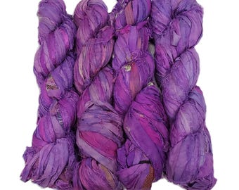 Può includere: Un fascio di filato di nastro di seta sari viola vibrante. Il filato è strettamente avvolto, mostrando sfumature di lavanda, viola e magenta. La trama appare morbida e leggermente lucida, ideale per vari progetti di artigianato come maglia, uncinetto e tessitura.