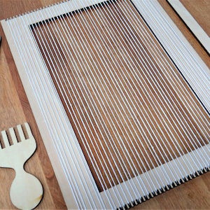 Op de afbeelding: Een houten weefgetouw met witte touw warp-draden, een houten kam en een houten naald. Het weefgetouw staat op een houten oppervlak.