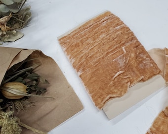 May include: A spool of light brown velvet ribbon wrapped around a rectangular card. The ribbon has a soft, textured surface with a slight sheen. The spool is positioned next to a brown paper bag containing dried flowers and foliage.