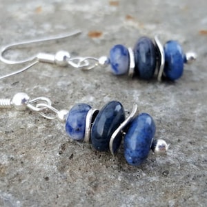May include: A pair of dangle earrings featuring silver-toned hooks and hardware. Each earring consists of three smooth, rounded blue sodalite beads separated by thin, curved silver-toned metal spacers, ending with a small silver-toned bead at the base.