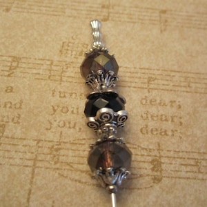 May include: A decorative pin with a silver-colored metal shaft and ornate top. The pin features dark brown and black faceted beads and decorative silver-colored metal accents. The background is a sheet of paper with musical notes and text.