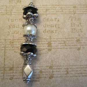 Peut inclure: Marque-page décoratif avec une base et des accents en métal argenté. Il présente des perles noires et transparentes, une fausse perle et des détails de filigrane ornés. Le fond est un motif de partition vintage.