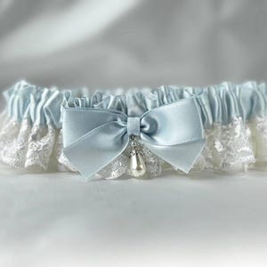 May include: A light blue satin garter featuring a ruffled design with white lace trim. The center is adorned with a matching light blue satin bow and a single teardrop-shaped faux pearl charm suspended from a small silver-toned metal cap.