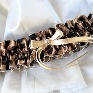 May include: A leopard print garter with a gold bow and pearl accent. The garter has a ruffled design with black and brown spots. Gold ribbon is attached to the bow. The garter is set against a white background.