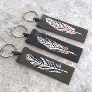 May include: Three brown leather keychains with silver key rings. Each keychain features a hand-painted white and gold feather design.