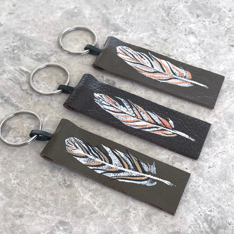 May include: Three brown leather keychains with silver key rings. Each keychain features a hand-painted white and gold feather design.