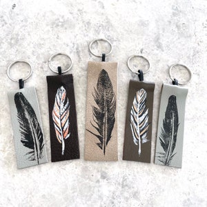May include: Five leather keychains with different feather designs. The keychains are in shades of brown, gray, and beige. Each keychain has a silver key ring.