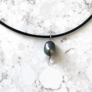 Puede incluir: Un collar gargantilla de cuero negro con un colgante de perla gris oscuro.