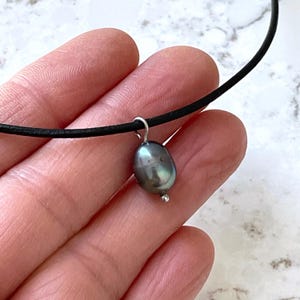 Puede incluir: Un collar de cord&oacute;n de cuero negro con un colgante de perla gris oscuro.