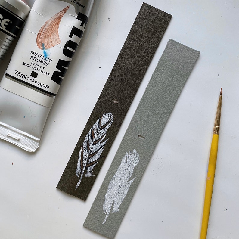 May include: Two leather strips with painted white feathers. The strips are brown and gray. A yellow paintbrush is in the background.