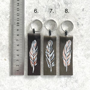 May include: Three leather keychains with silver key rings. Each keychain features a painted feather design in white, brown, and gold on a dark green background. The keychains are numbered 6, 7, and 8.