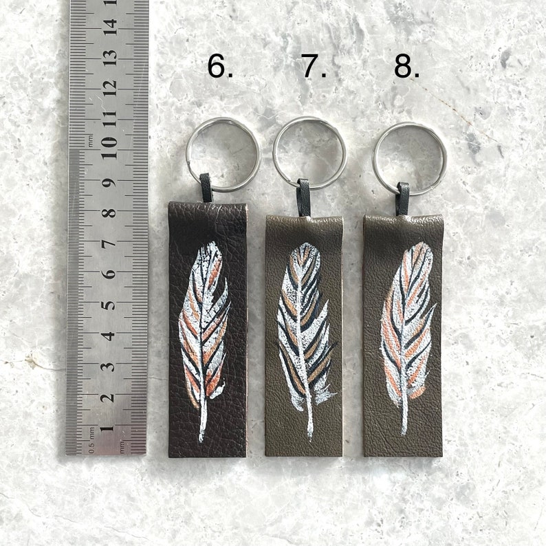 May include: Three leather keychains with silver key rings. Each keychain features a painted feather design in white, brown, and gold on a dark green background. The keychains are numbered 6, 7, and 8.