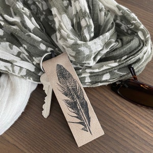May include: A brown leather keychain with a black feather design. The keychain is attached to a set of keys and a gray and white patterned scarf.