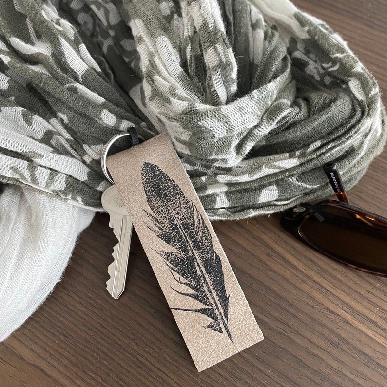 May include: A brown leather keychain with a black feather design. The keychain is attached to a set of keys and a gray and white patterned scarf.